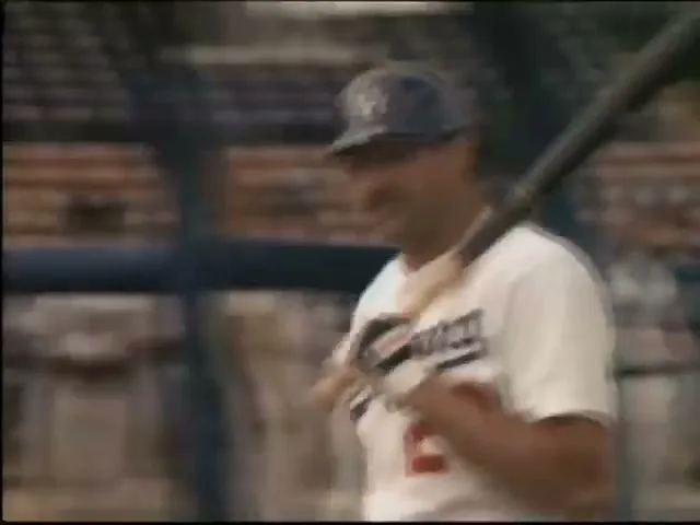 A baseball player in a white uniform and blue cap stands on a baseball field, holding a bat over his shoulder, looking serious.