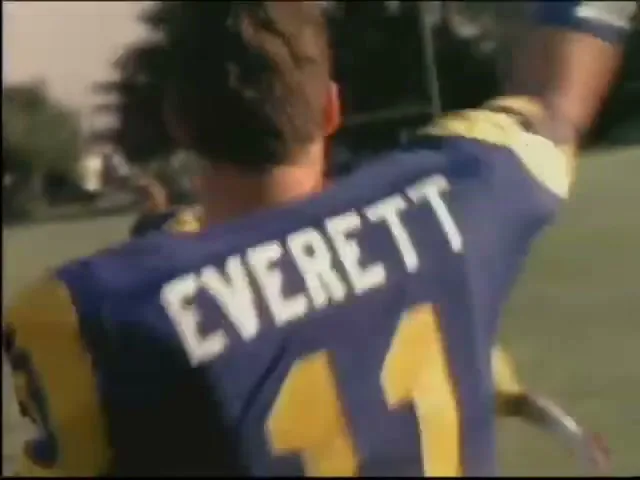 A football player in a blue and yellow jersey with the name "EVERETT" and number "11" on the back, turns to face the camera on a sunny football field.