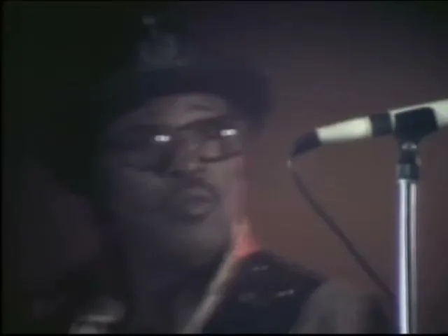 A man in a hat and sunglasses sings into a microphone on stage, then another man in a tank top appears, looking up.