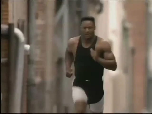 Bo Jackson, a muscular Black man, runs towards the camera in a black tank top and white shorts down a narrow urban alleyway during the day.
