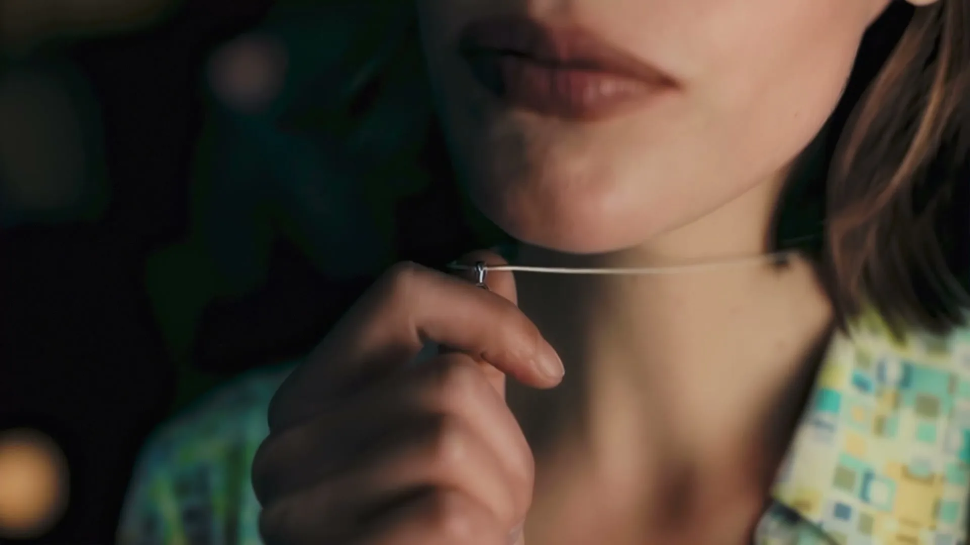 A young woman with light brown hair and a patterned shirt adjusts a small pendant on a thin necklace around her neck.