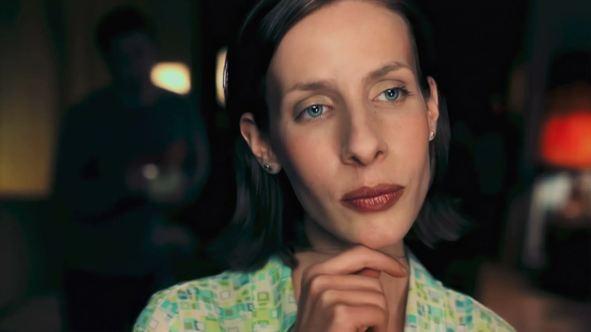 A woman with blue eyes and short brown hair, wearing a patterned shirt, looks thoughtfully to the side in a dimly lit room.