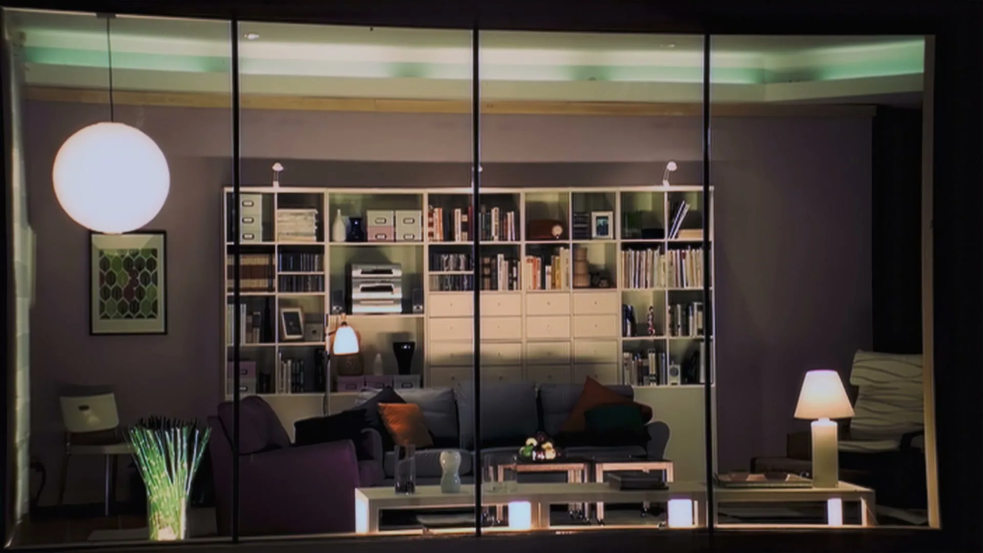 A wide shot of a modern living room with a large bookshelf, sofa, and various furniture pieces, viewed through a window at night.