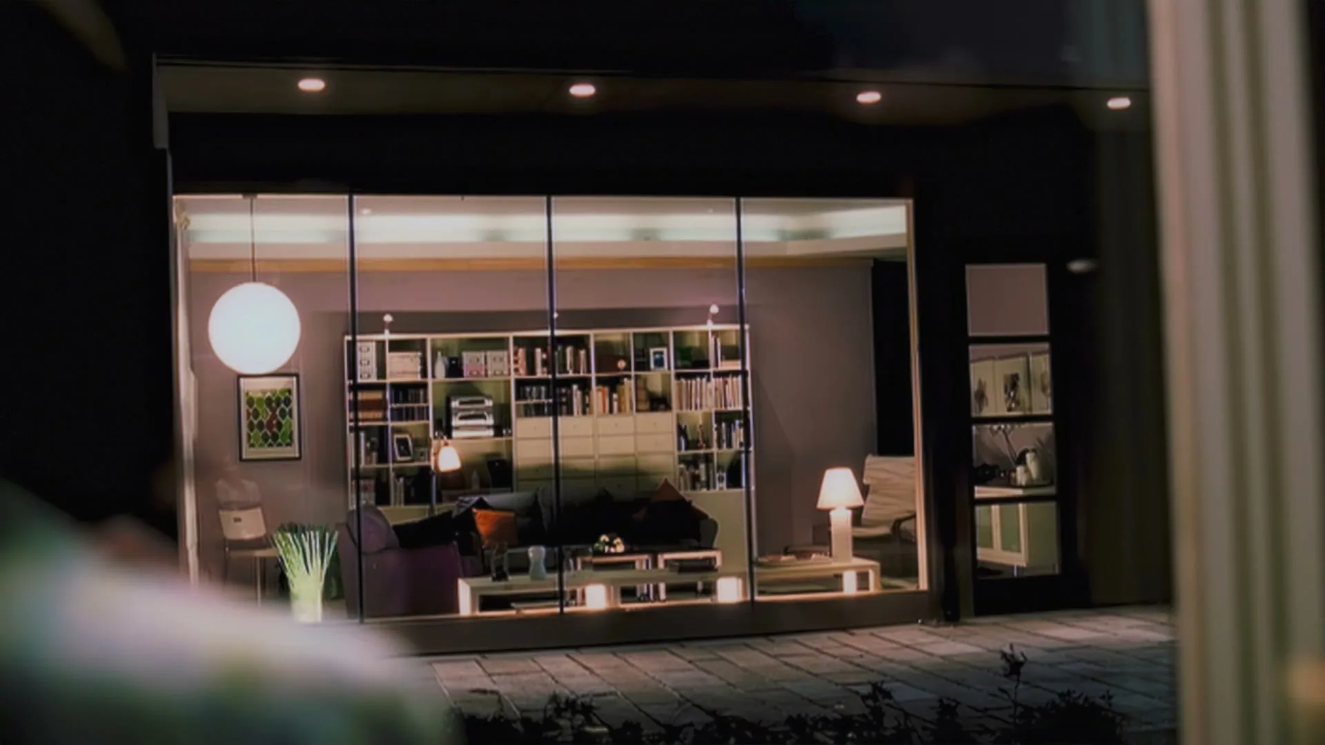 A wide shot of a modern living room with a bookshelf and sofa, seen through a large window at night.