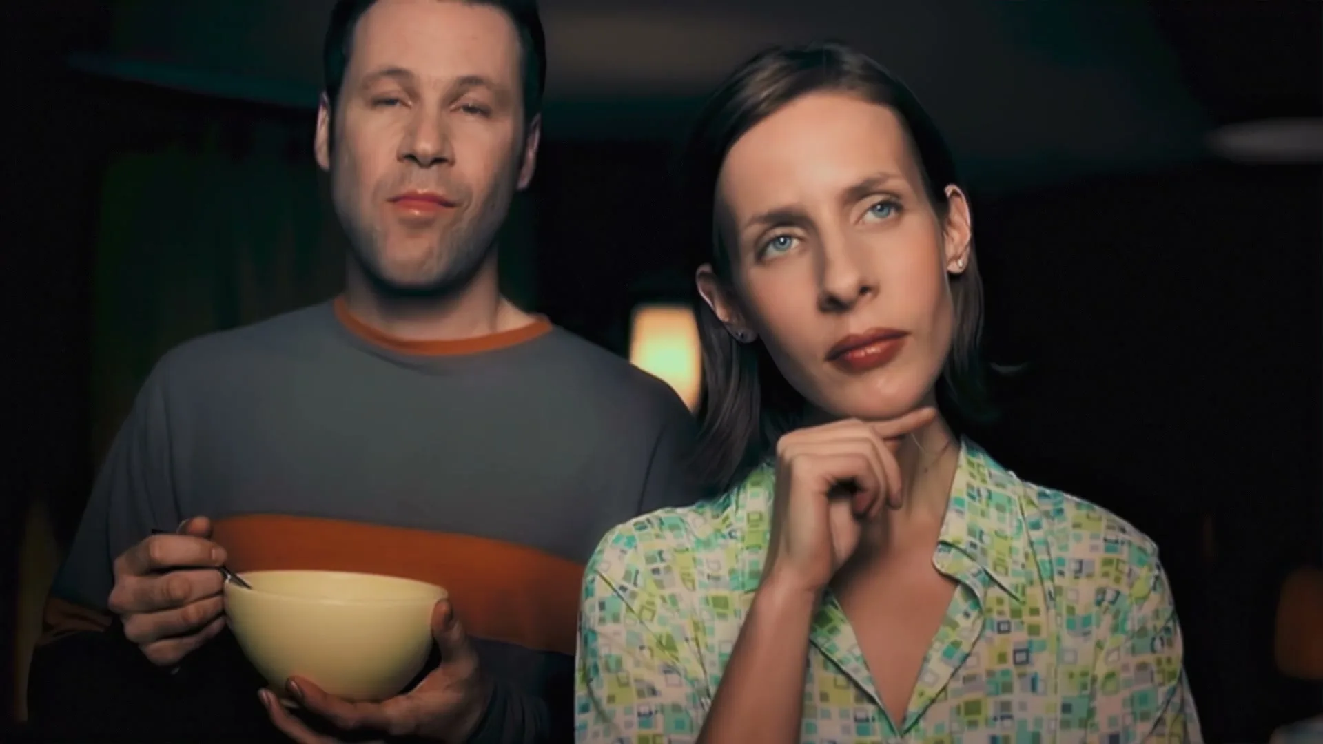 A man in a grey and orange shirt holds a bowl while a woman in a patterned shirt looks thoughtful, both standing in a dimly lit interior.
