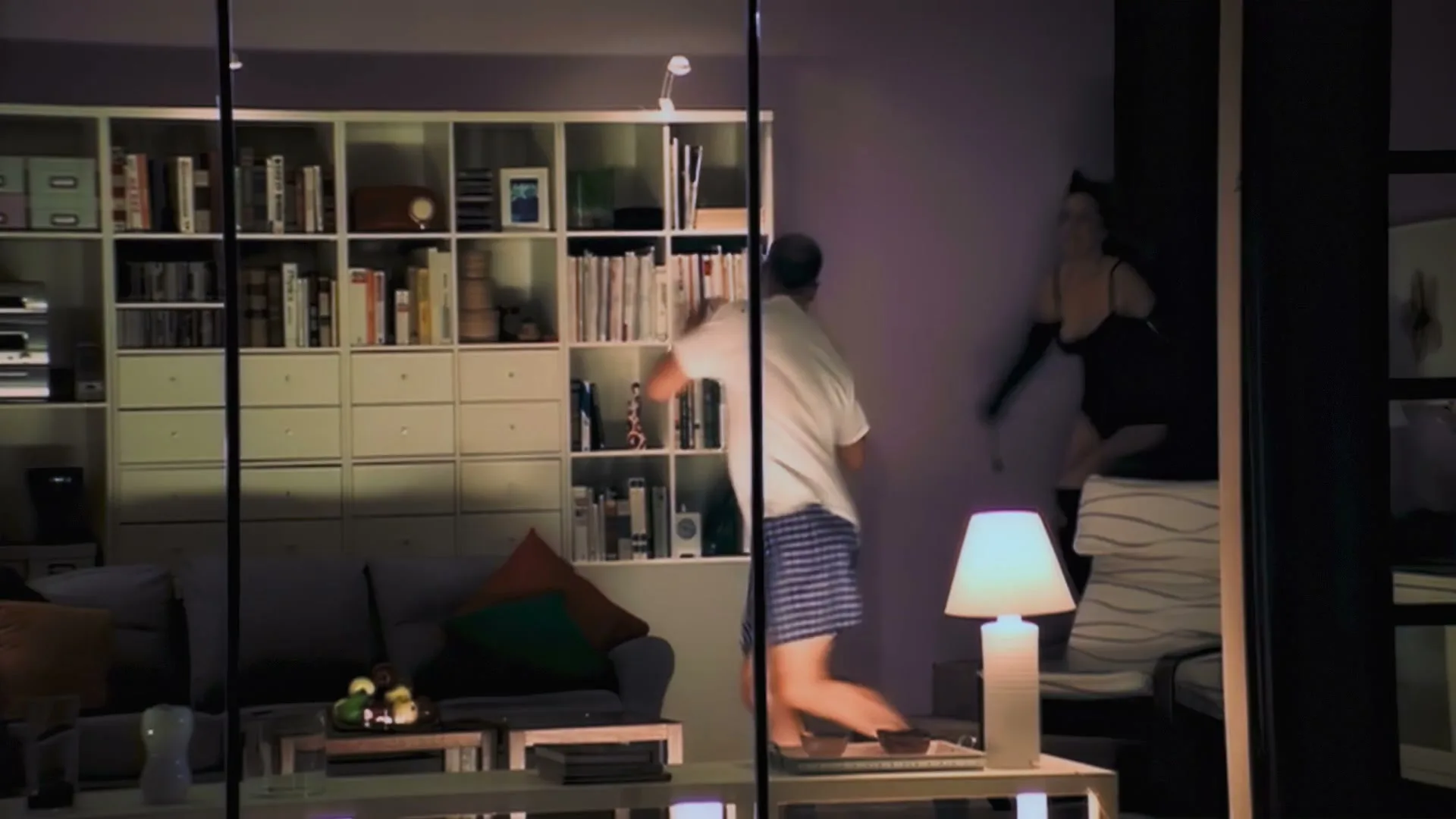 A man in boxers and a woman in lingerie are seen through a window, as the man points at the woman in a living room with a large bookshelf.
