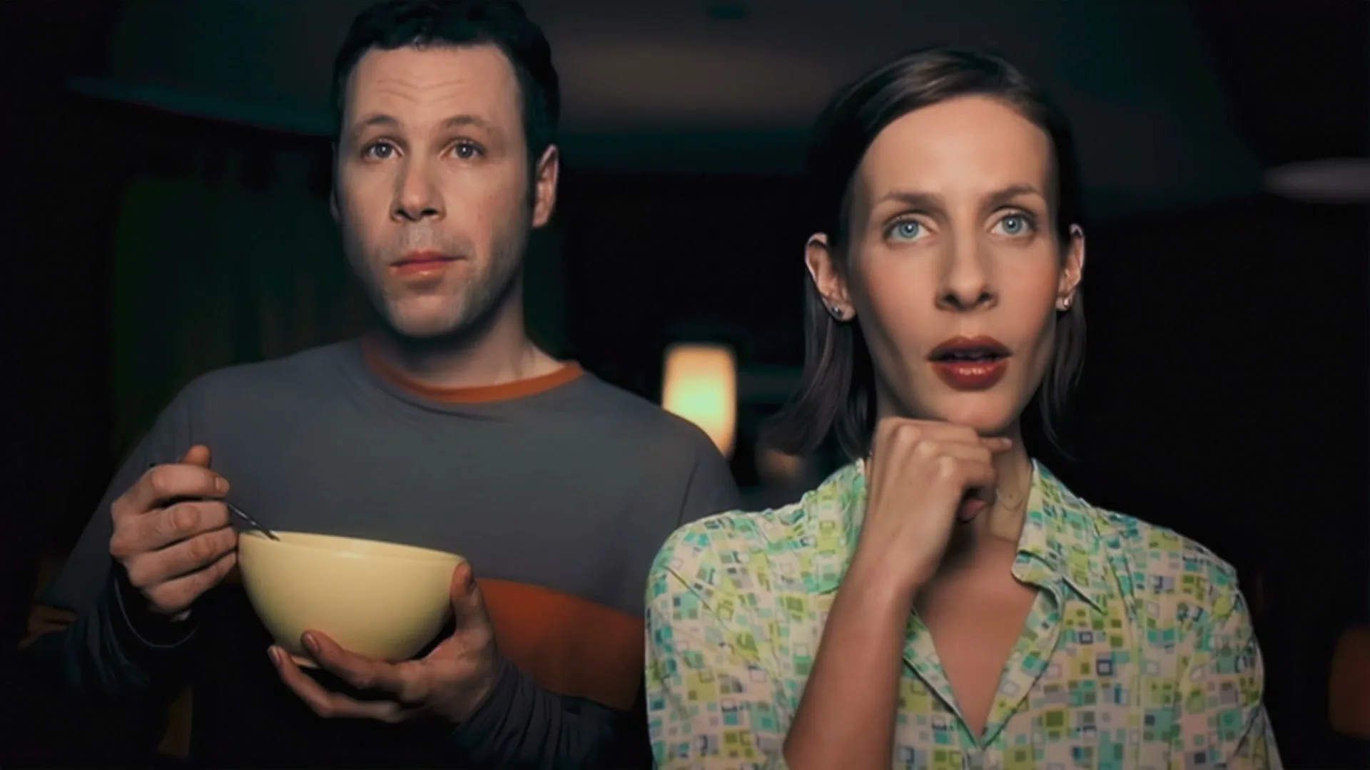 A man and woman sit side-by-side, looking forward with serious expressions, as the man holds a bowl and spoon in an interior setting.