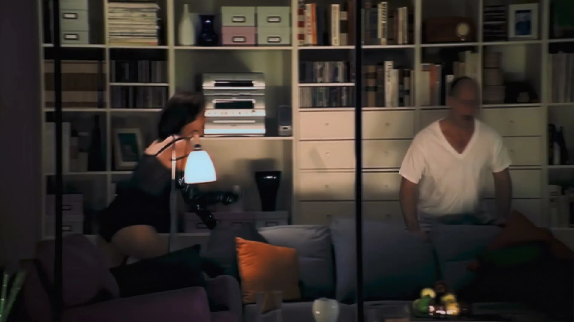 A man and woman in a living room, the woman in lingerie, the man in a t-shirt, as they move around a sofa in front of a large bookshelf.