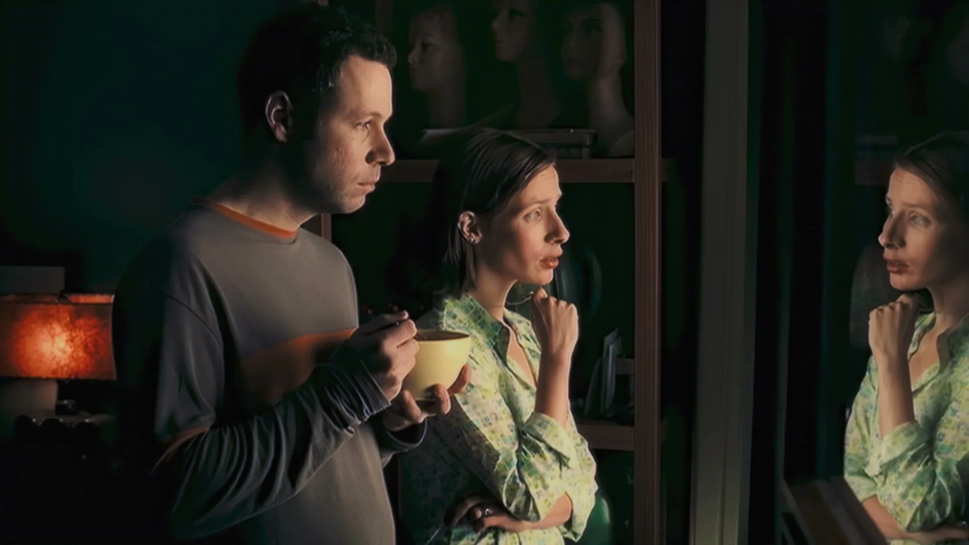 A man and a woman in pajamas stand in a dimly lit room, looking at the woman's reflection in a mirror, with mannequin heads on a shelf behind them.