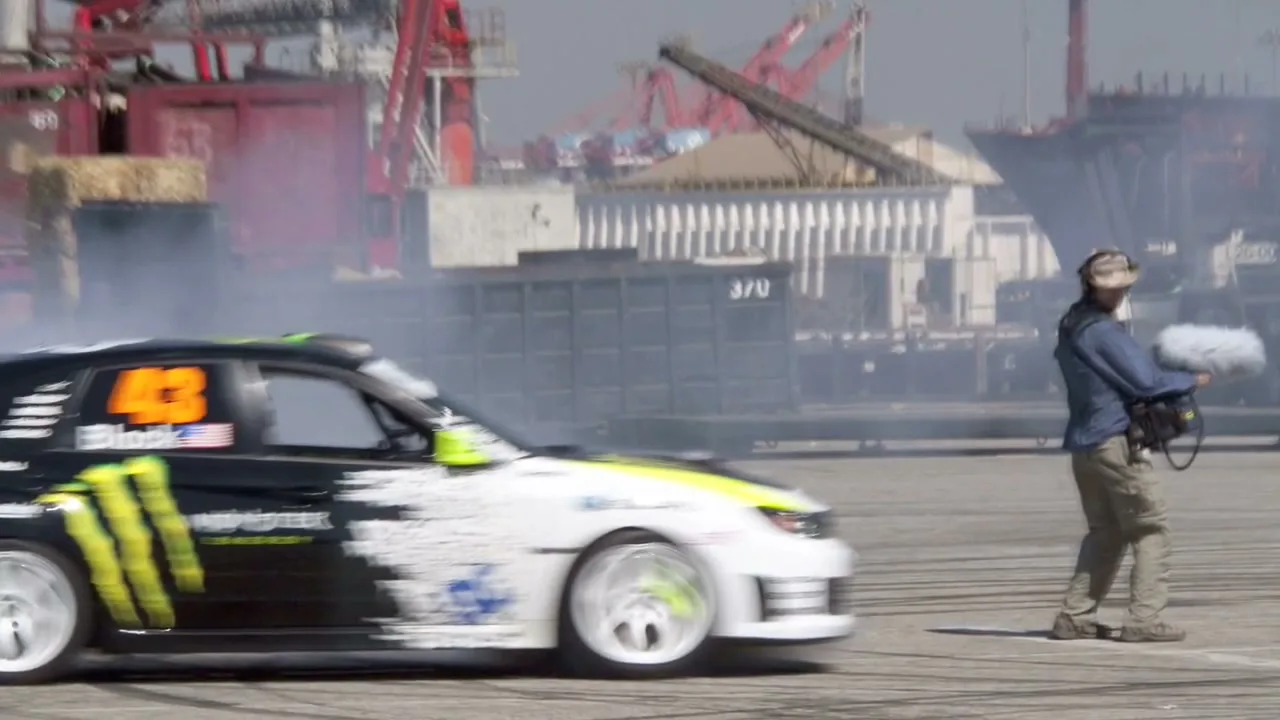 A sound engineer with a boom mic records a black and white rally car with green accents drifting and smoking in an industrial port area during the day.