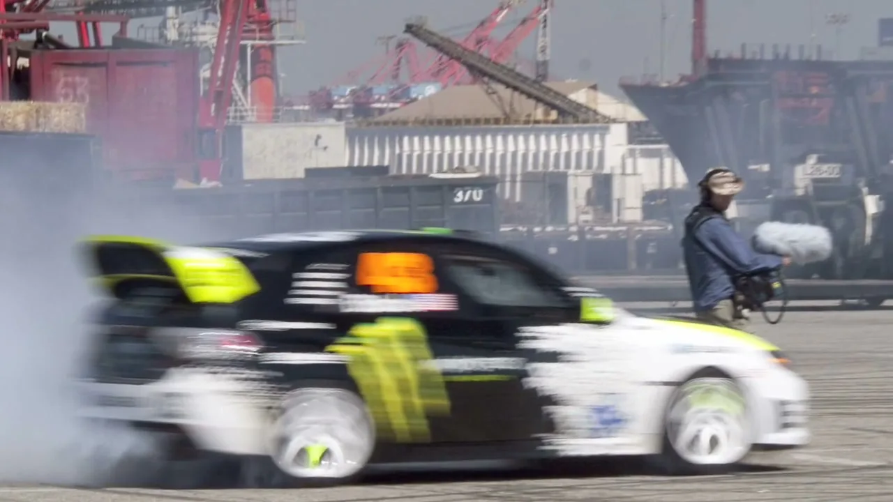 A black and white rally car with green accents drifts, creating a cloud of smoke, while a cameraman films in an industrial port.