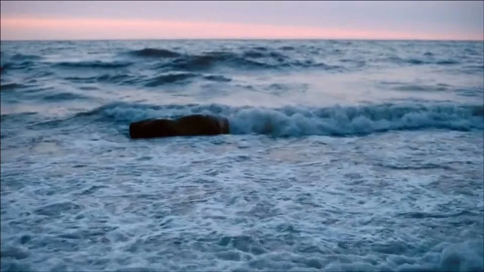 The ocean waves crash against rocks at dusk, with a soft pink and purple sky in the background.
