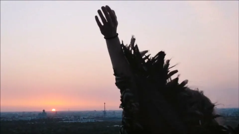 A person with long hair and a feathered garment raises their arm towards a sunset over a city skyline and forest.