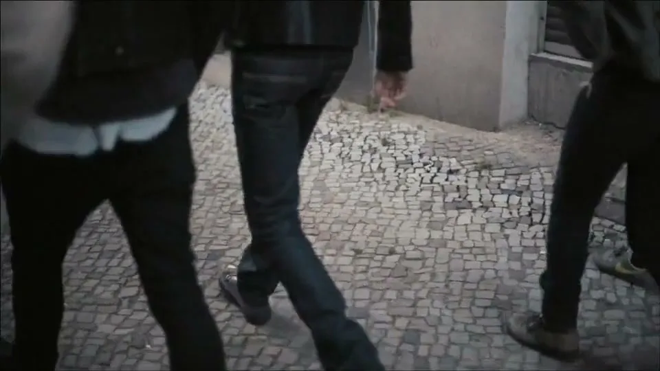 Three people walk away from the camera on a cobblestone street, wearing dark clothing and jeans, in an urban setting.