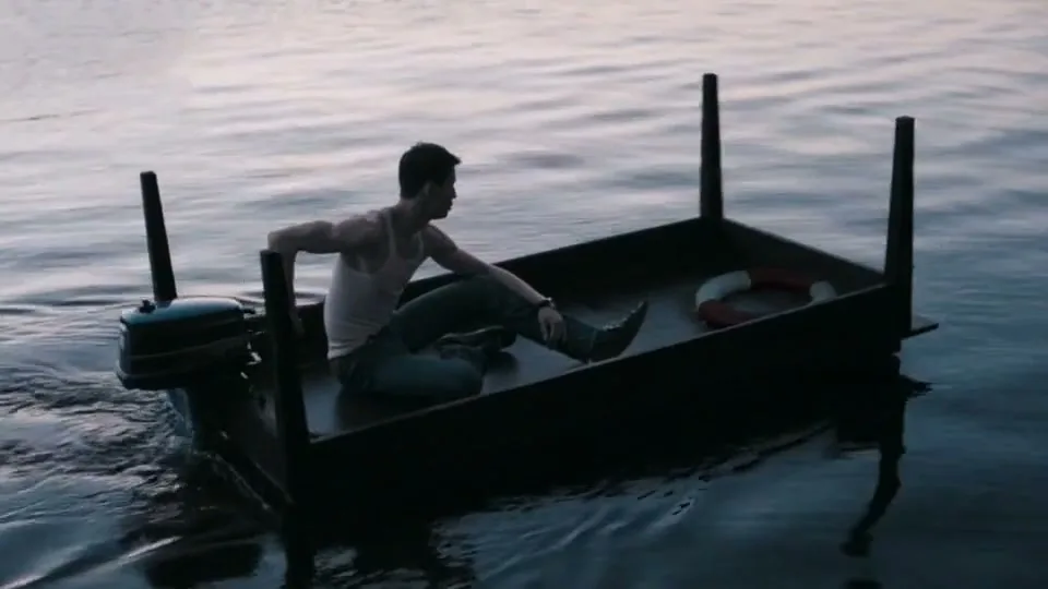 A young man in a tank top and jeans sits in a makeshift boat made from a bed frame, powered by an outboard motor, on calm water.
