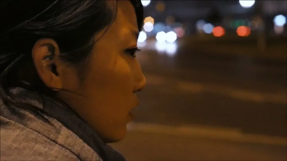 A young woman with dark hair and a grey hoodie looks off-screen to the right, with blurred city lights in the background at night.