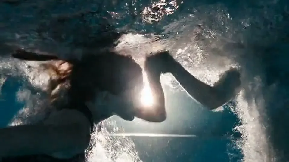 A young woman with long hair swims underwater in a pool, with sunlight dappling through the surface, creating a serene and ethereal atmosphere.