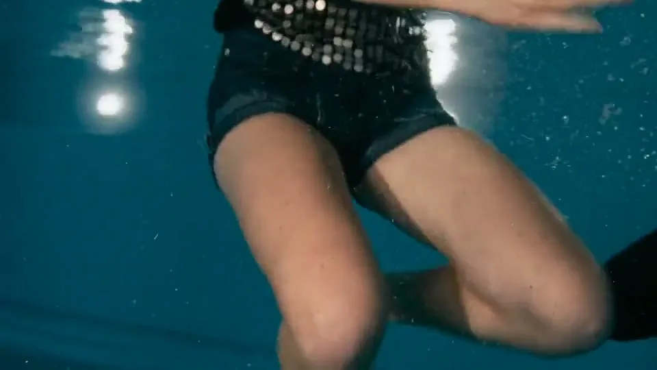 A young woman in a sequined top and denim shorts swims underwater in a pool, with light reflecting on the surface.