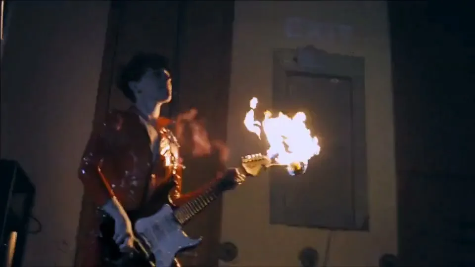 A young male musician in a shiny red jacket plays a flaming electric guitar in a dark, industrial-looking room with an exit sign.