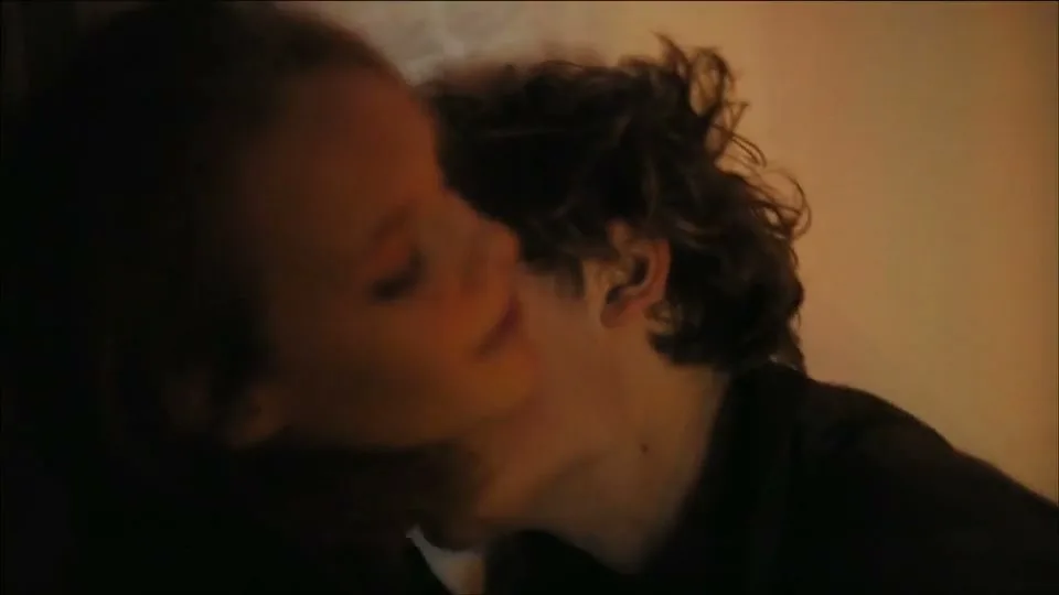 A young couple, a female with brown hair and a male with curly brown hair, kiss passionately in a dimly lit room.