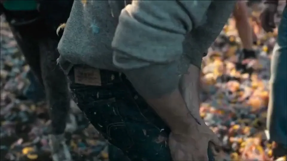 A person in a grey hoodie and blue jeans is seen from the waist down, bending over in a pile of colorful leaves.