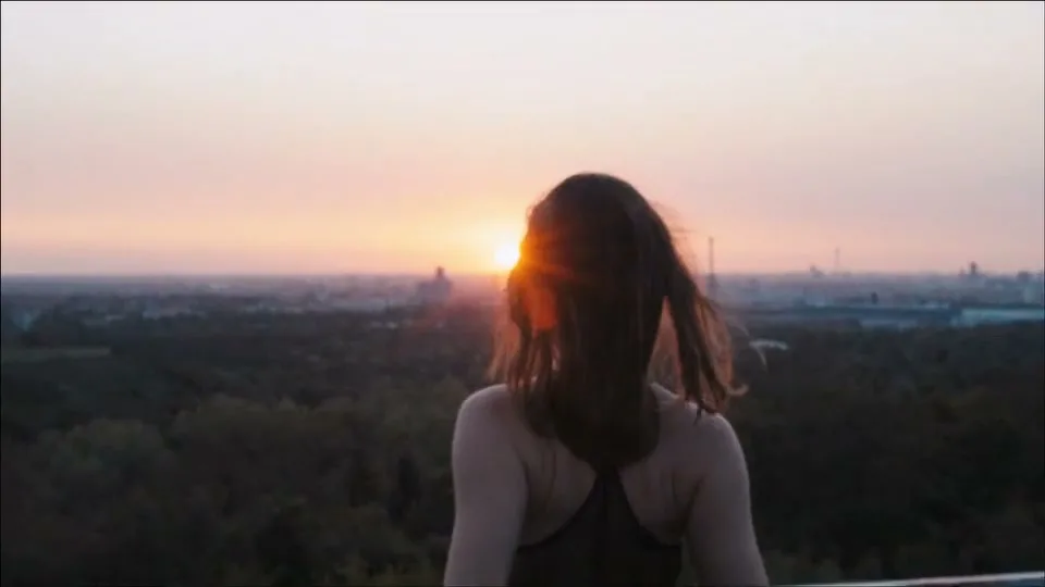 A young woman with long brown hair, seen from behind, stands on a rooftop overlooking a city skyline as the sun sets.