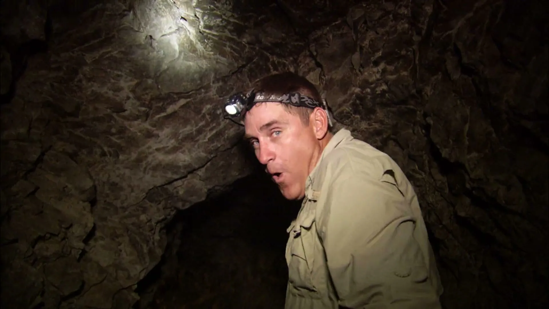 A man wearing a headlamp and khaki shirt looks around a dark cave with a surprised expression, illuminated by his headlamp.
