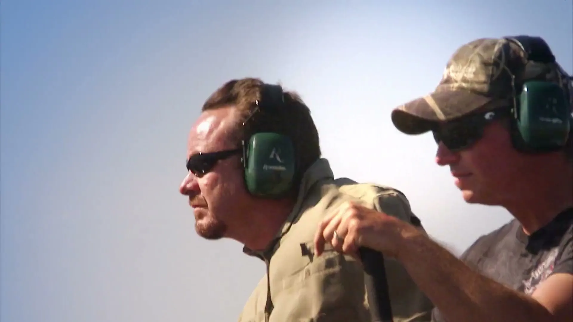 Two men wearing ear protection and sunglasses look intently to the left, one with a hand on the other's shoulder, against a bright, hazy sky.
