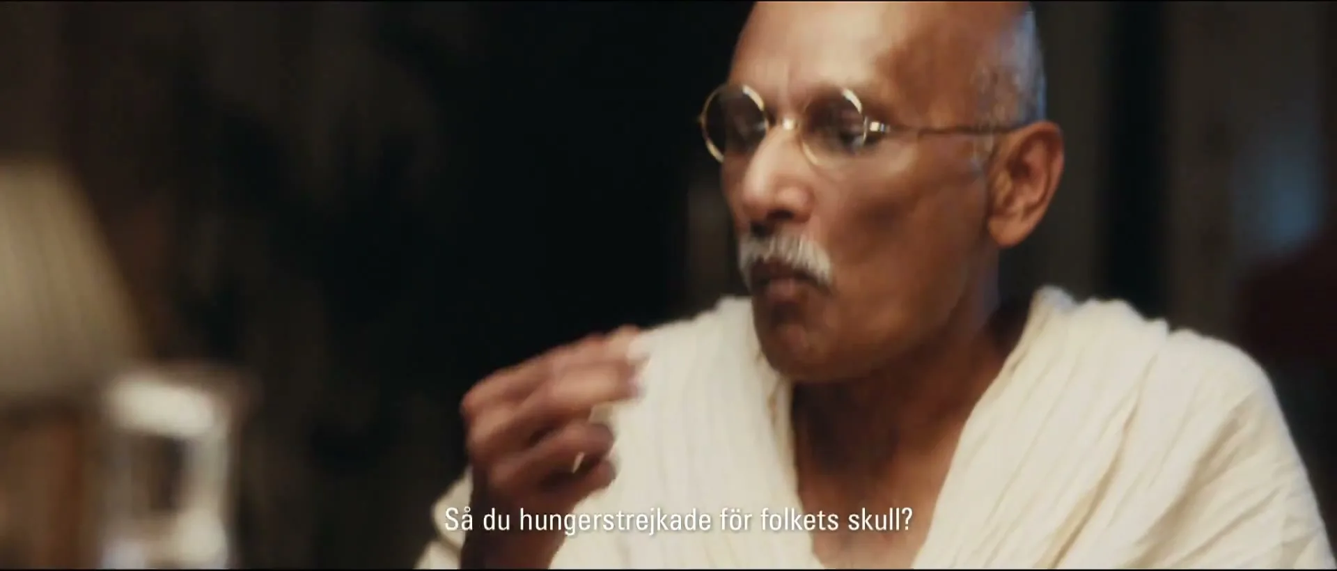 A man resembling Mahatma Gandhi, with a bald head, round glasses, and a white mustache, sits at a table, eating grapes from a small plate.