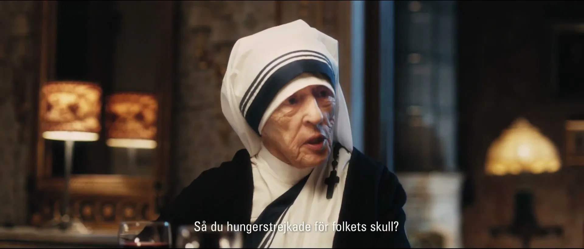 An elderly woman dressed as Mother Teresa sits at a dining table, speaking with a serious expression in a dimly lit room.