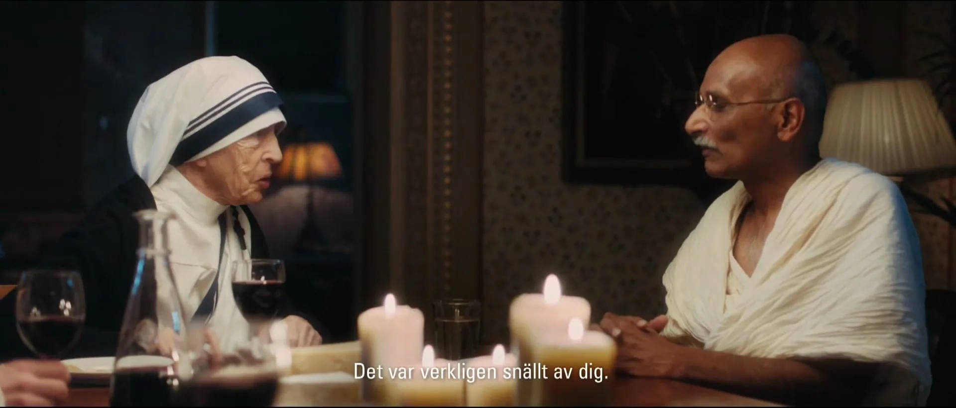 Mother Teresa and Mahatma Gandhi, portrayed by actors, sit at a candlelit dinner table, engaged in conversation in a dimly lit room.