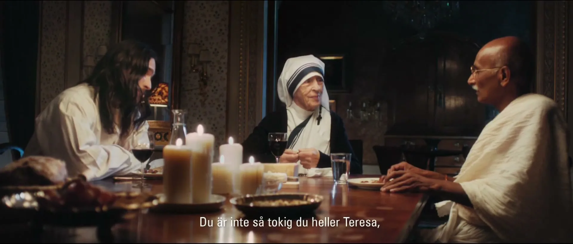 Three historical figures, including Mother Teresa, sit around a candlelit dining table, engaged in conversation during a Christmas dinner.