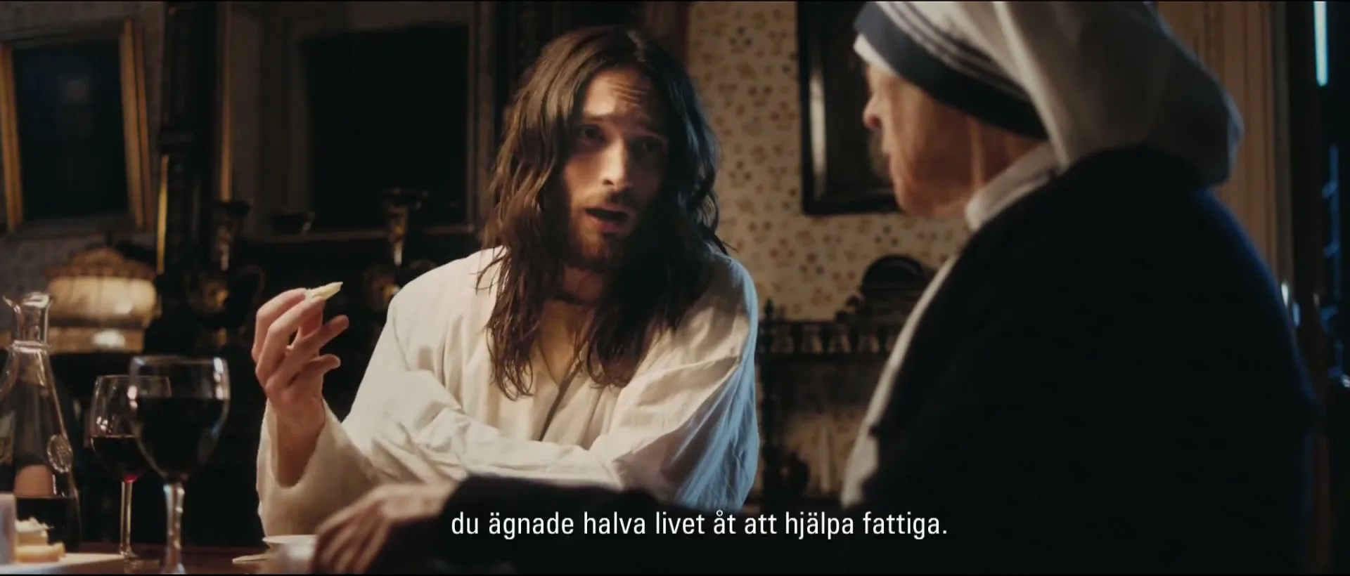 A man resembling Jesus, with long brown hair and a white robe, sits at a table, holding a piece of food and talking to an older woman dressed as Mother Teresa.