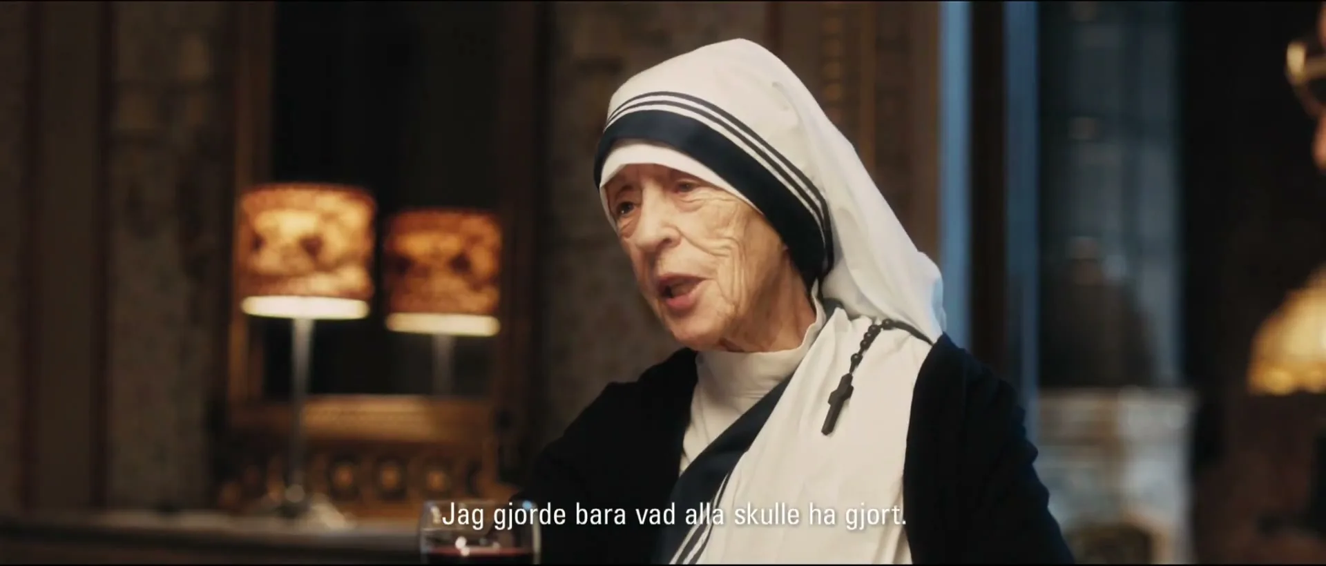 An elderly woman dressed as Mother Teresa speaks to an unseen person at a formal dinner table in a dimly lit room.