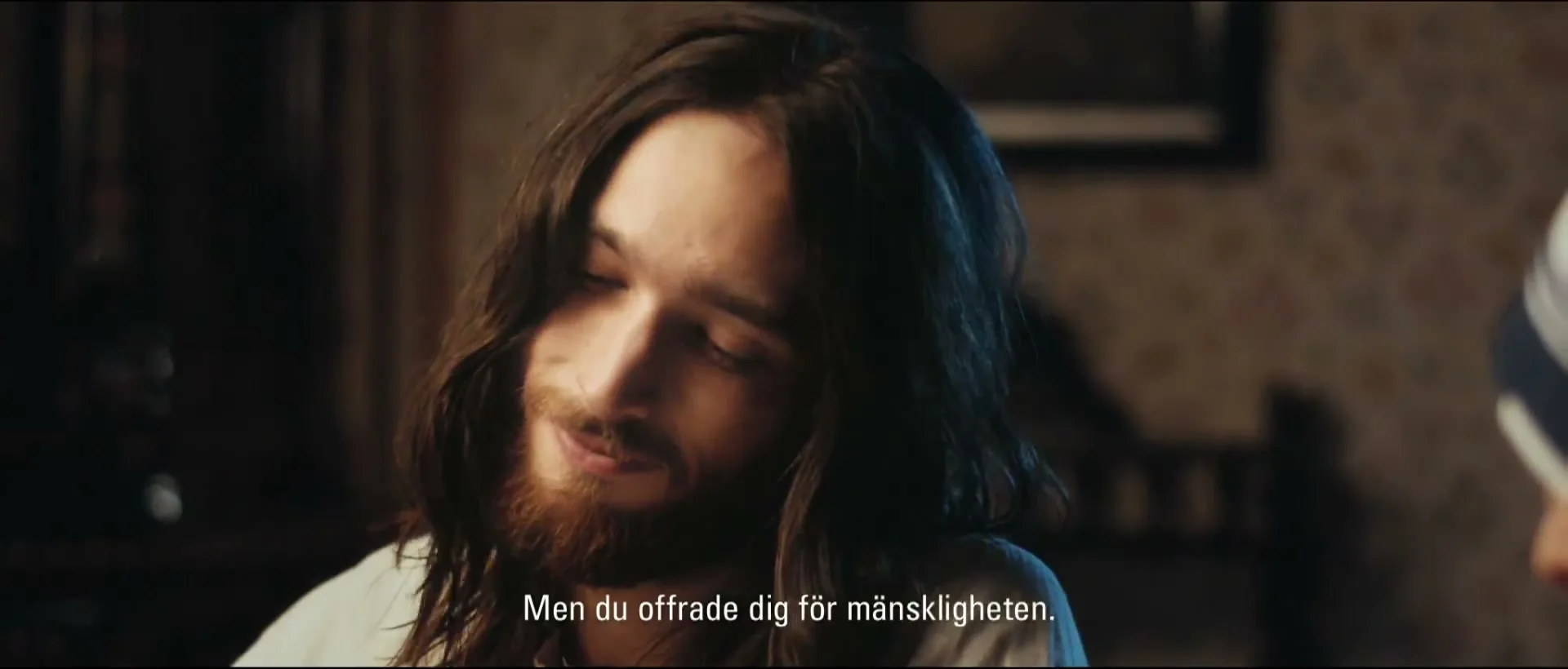 A young man with long hair and a beard looks down with a gentle smile, listening intently in a dimly lit room.