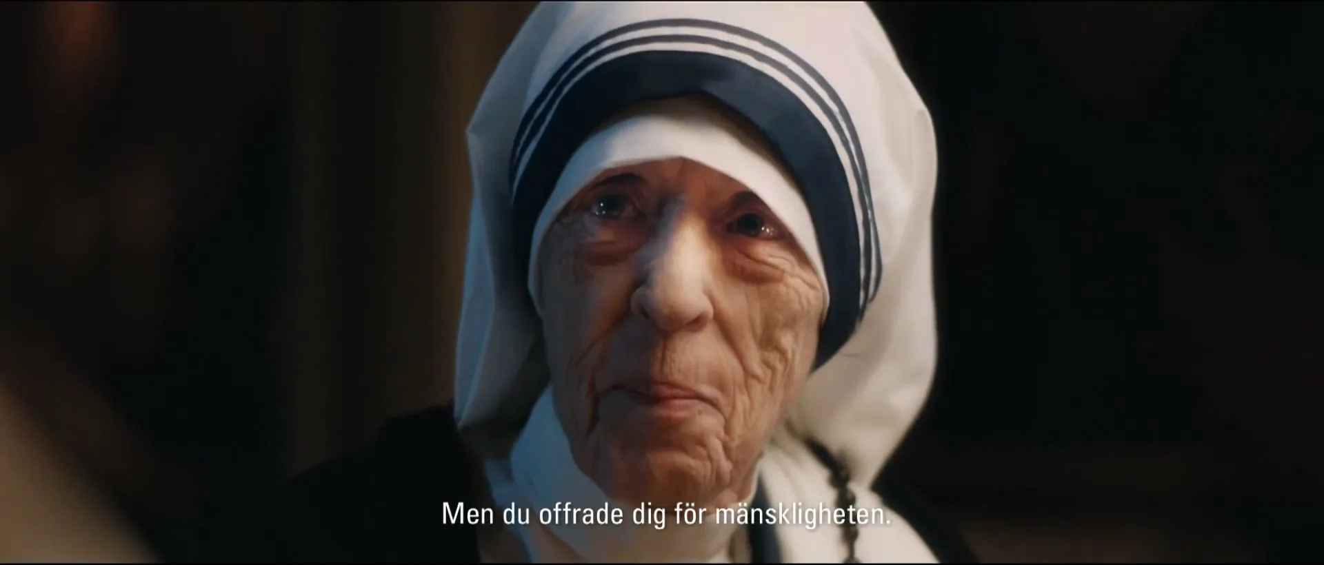 An elderly woman, resembling Mother Teresa, with a white habit and blue stripes, speaks directly to the camera in a dimly lit interior.