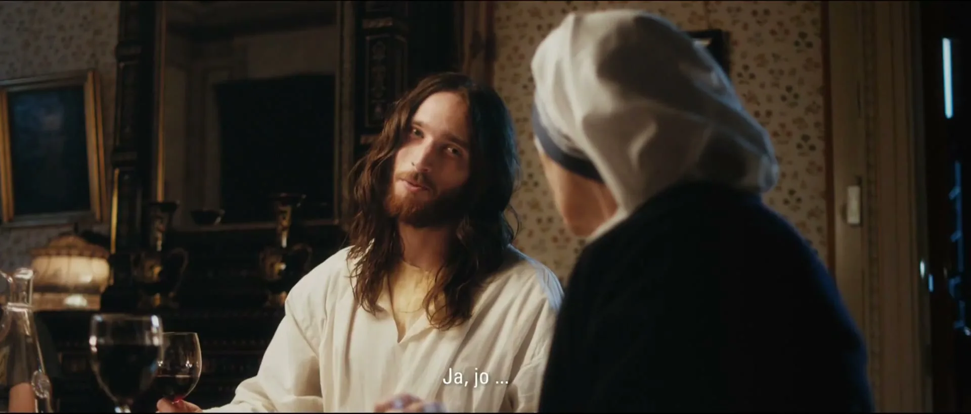 A man with long hair and a beard, dressed in a white tunic, sits at a table with a woman in a nun's habit, looking at her with a slight smile.