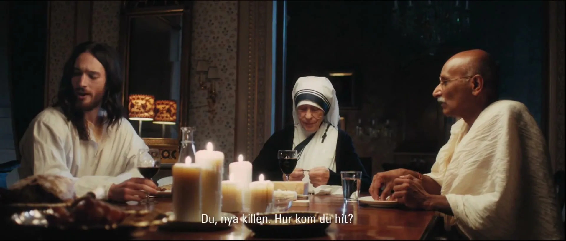Jesus, Mother Teresa, and Gandhi sit around a candlelit dining table, looking at someone off-screen during a meal.