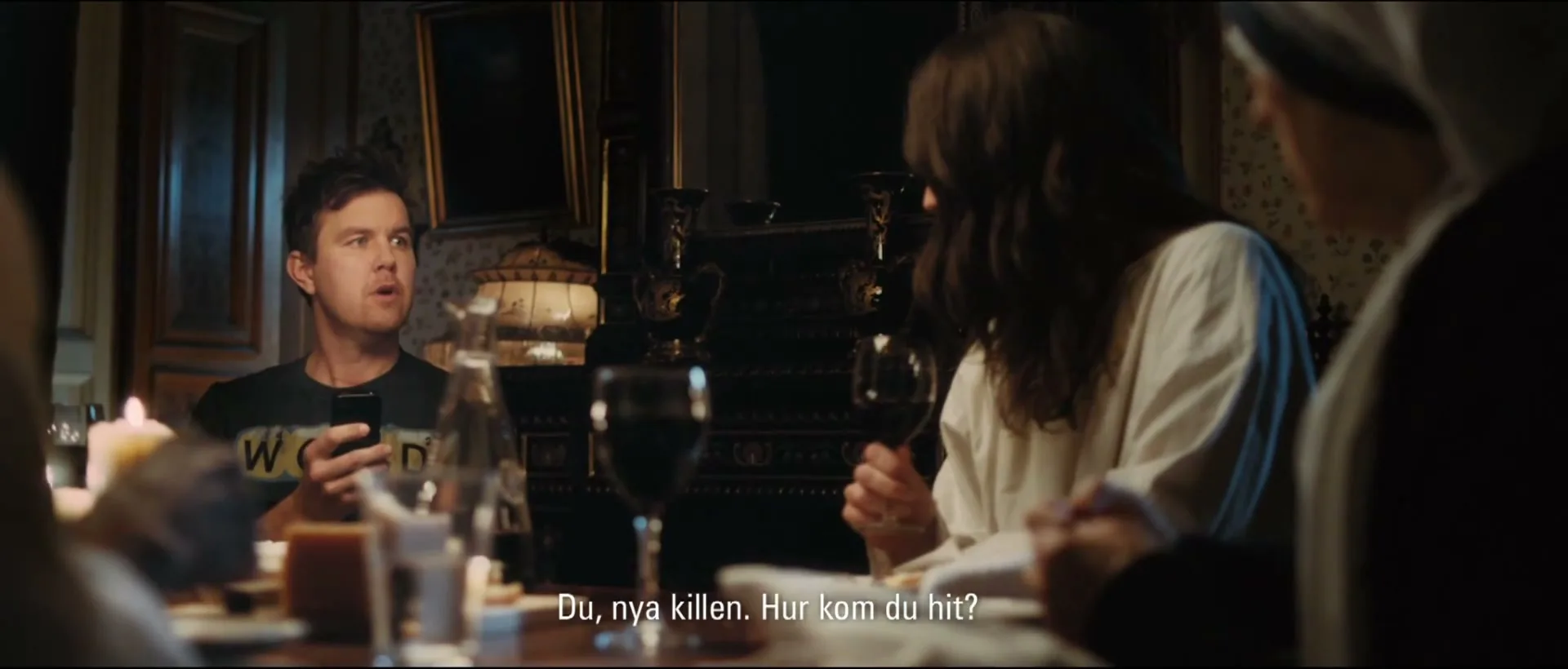 A man in a black t-shirt looks up from his phone at a dinner table, while a woman in a white robe holds a wine glass, in a dimly lit room.