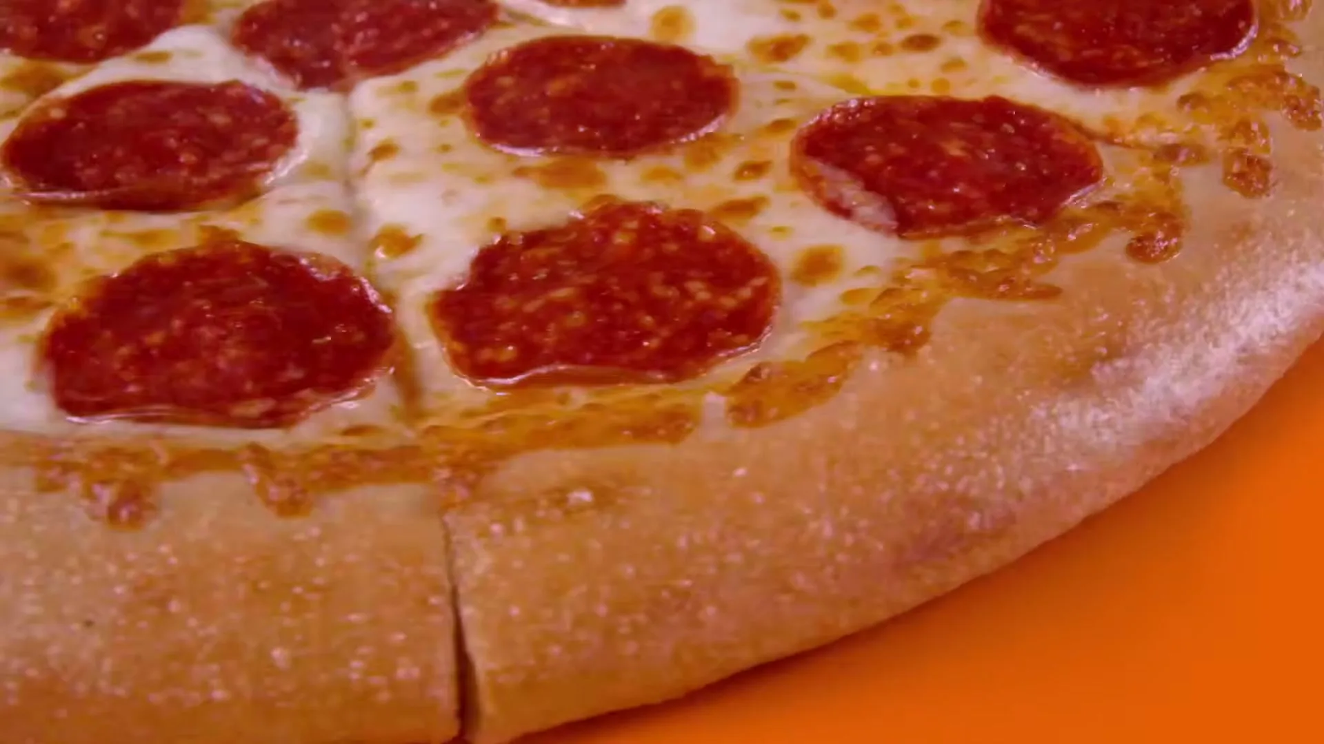 A whole pepperoni pizza with melted cheese is shown on an orange background, with the camera zooming in on the crust.
