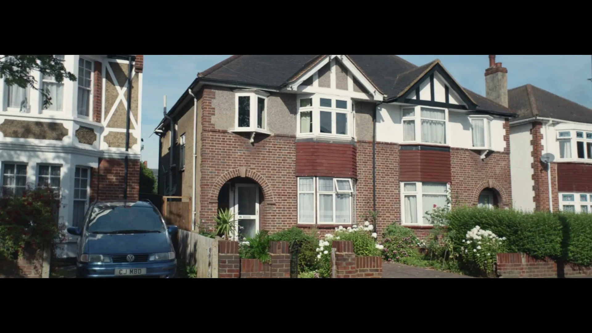 A wide shot of a suburban street with a blue car parked in front of a brick house with white trim and a garden.