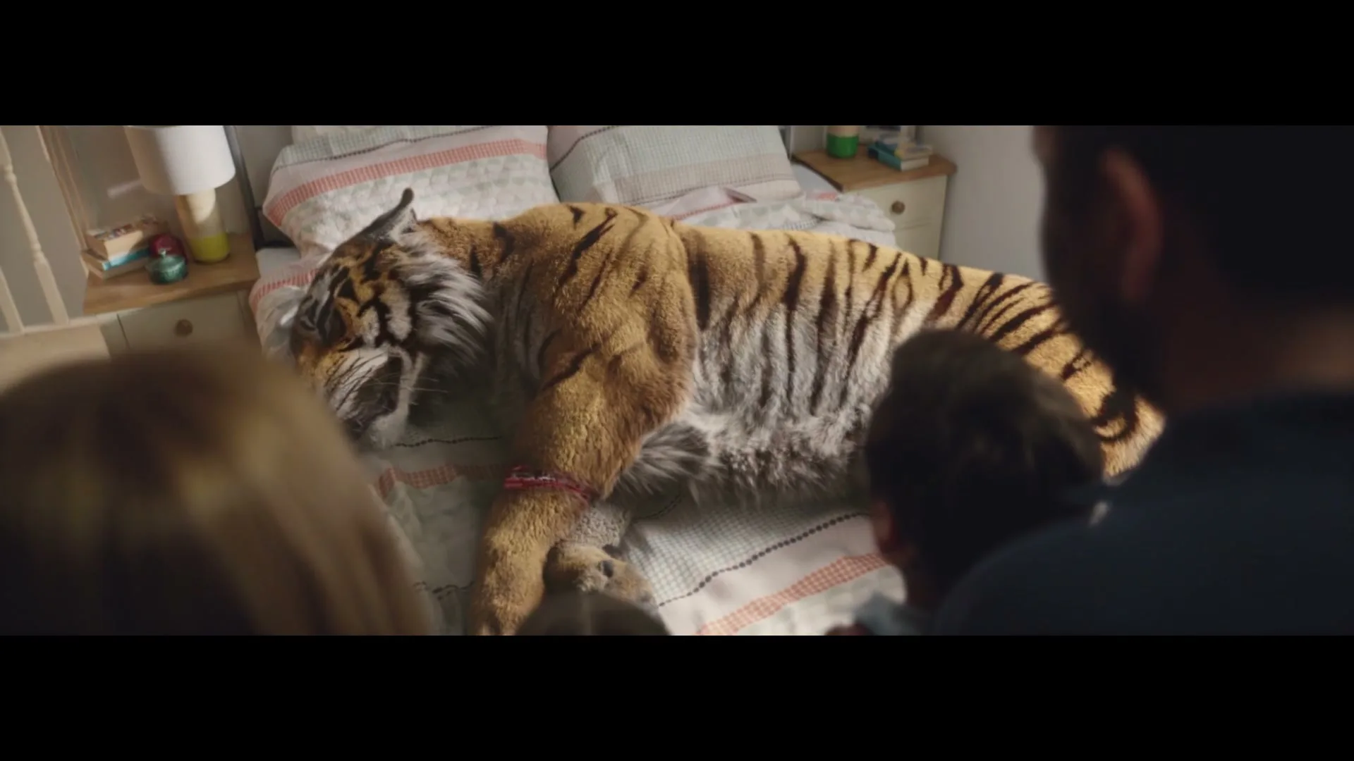 A family of four, seen from behind, looks at a large tiger sleeping on a bed in a child's bedroom.