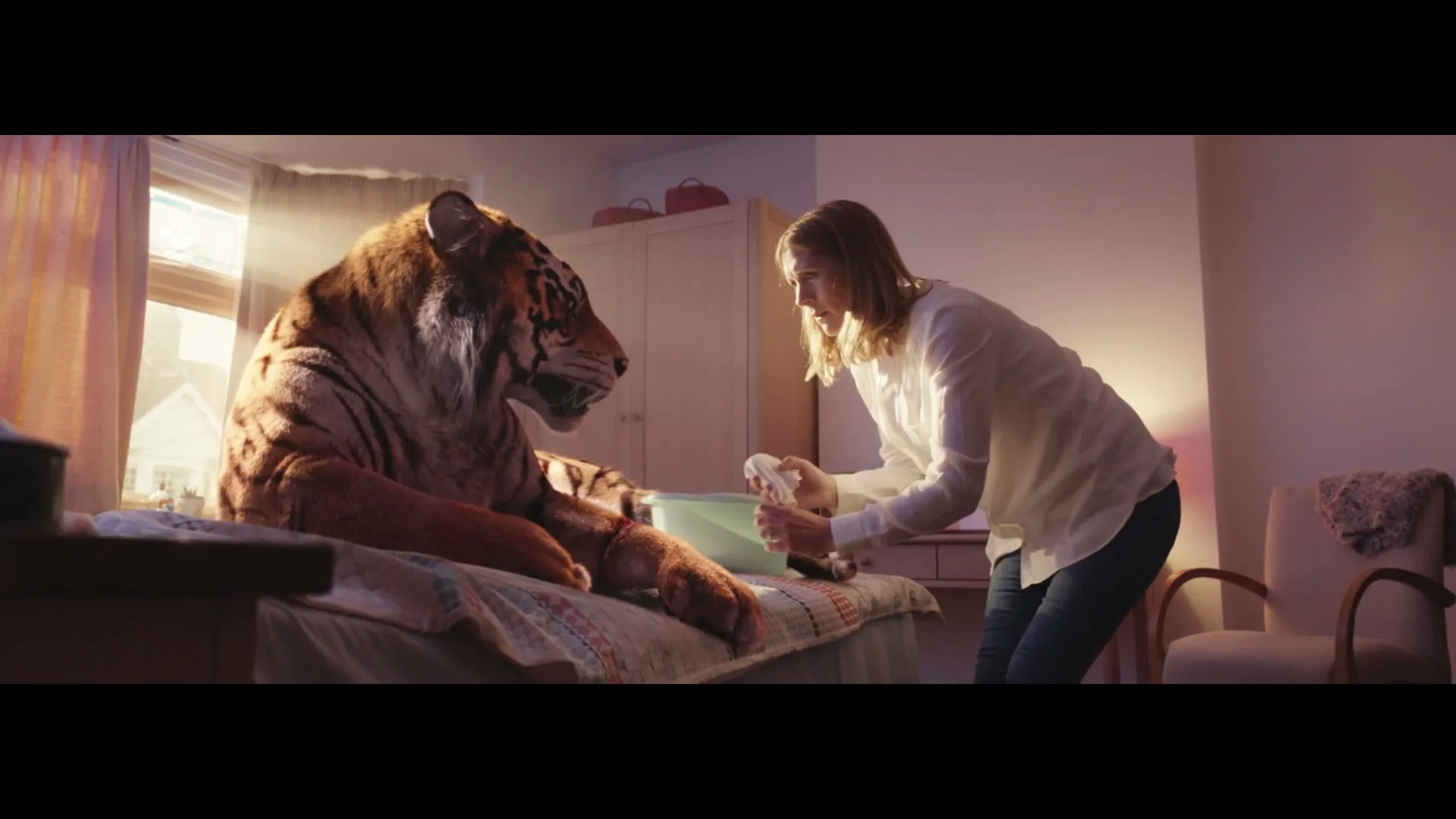 A woman in a white shirt and jeans tends to a large tiger lying on a bed in a sunlit bedroom.