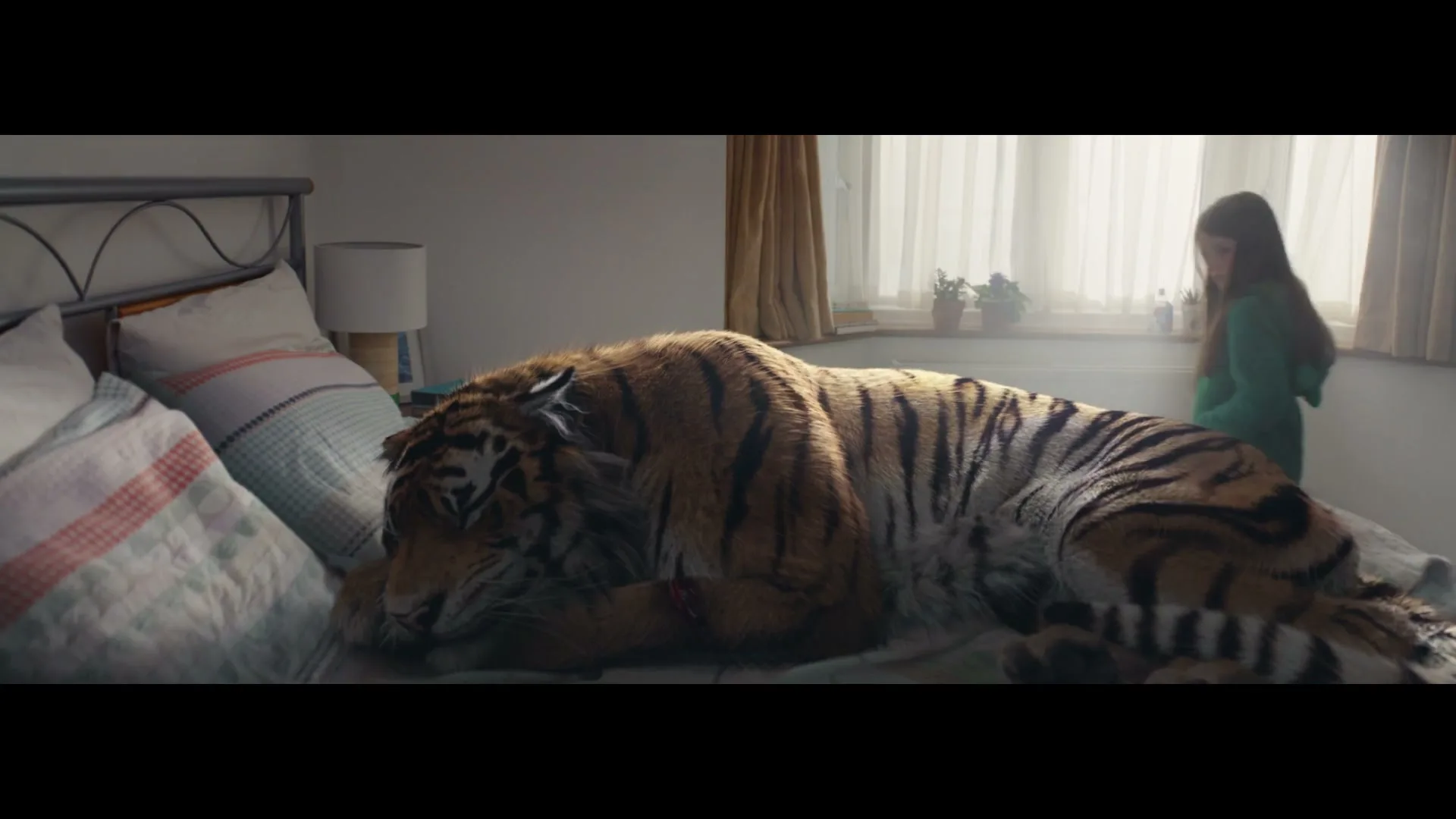 A young girl in a green hoodie stands by a window, looking at a large tiger sleeping on a bed in a sunlit bedroom.