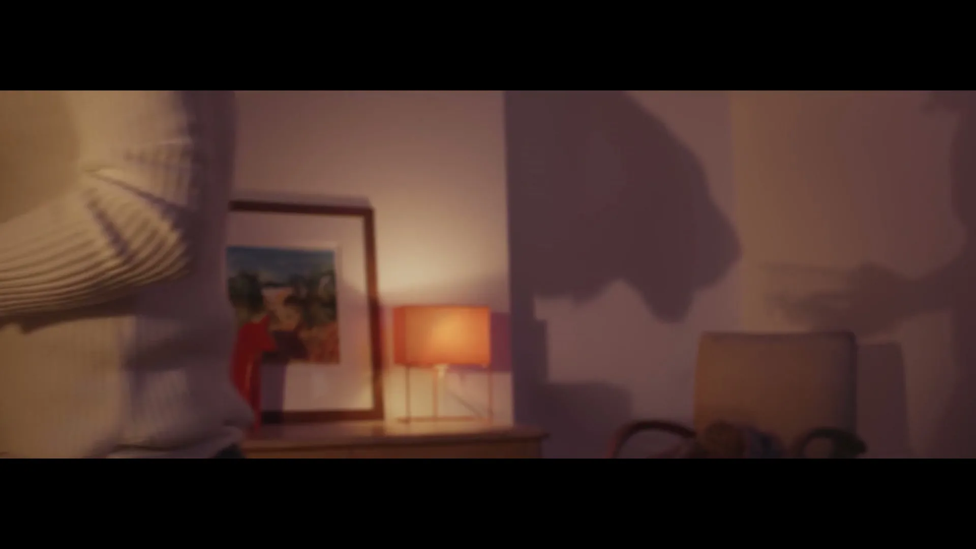 A woman in a white sweater walks past a dresser with a lamp and a framed picture, as a large shadow of a tiger appears on the wall.