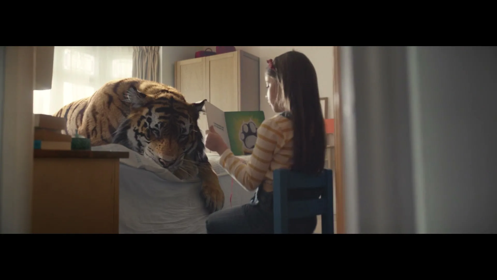 A young girl sits on a chair, reading a book to a large tiger lying on a bed in a sunlit bedroom.