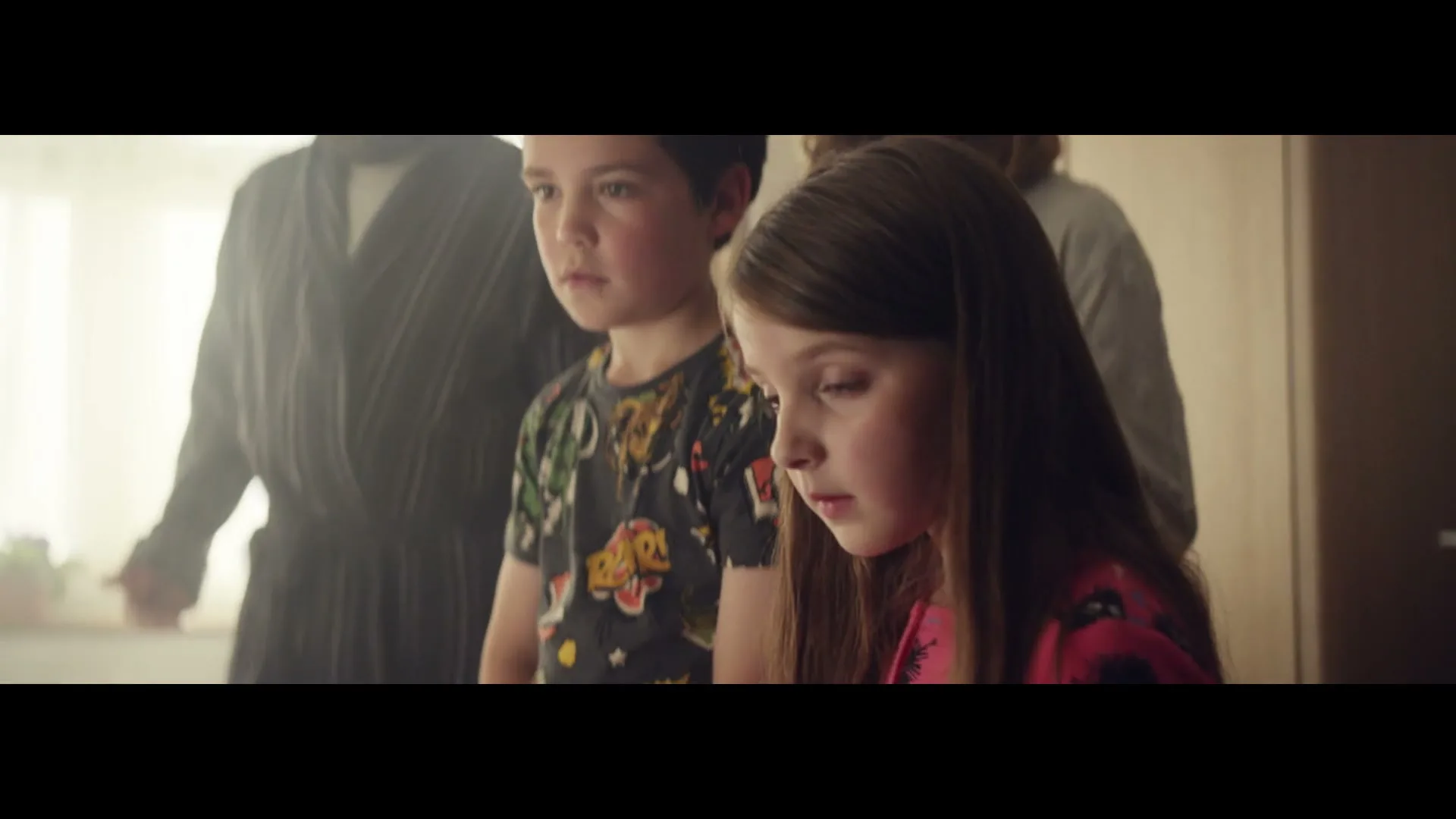 A young boy and girl stand in the foreground, looking down with serious expressions, while adults are blurred in the background.