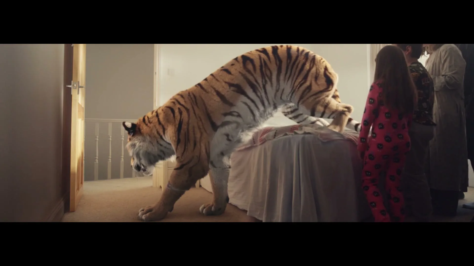A large, realistic tiger with orange and black stripes climbs onto a bed in a bedroom, while a small group of people watch in awe.