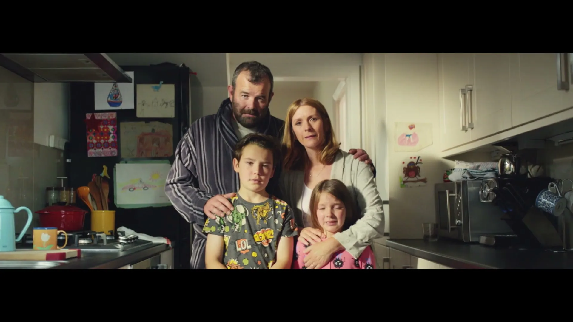 A family of four, a man, woman, and two children, stand together in a kitchen, looking directly at the camera with somber expressions.
