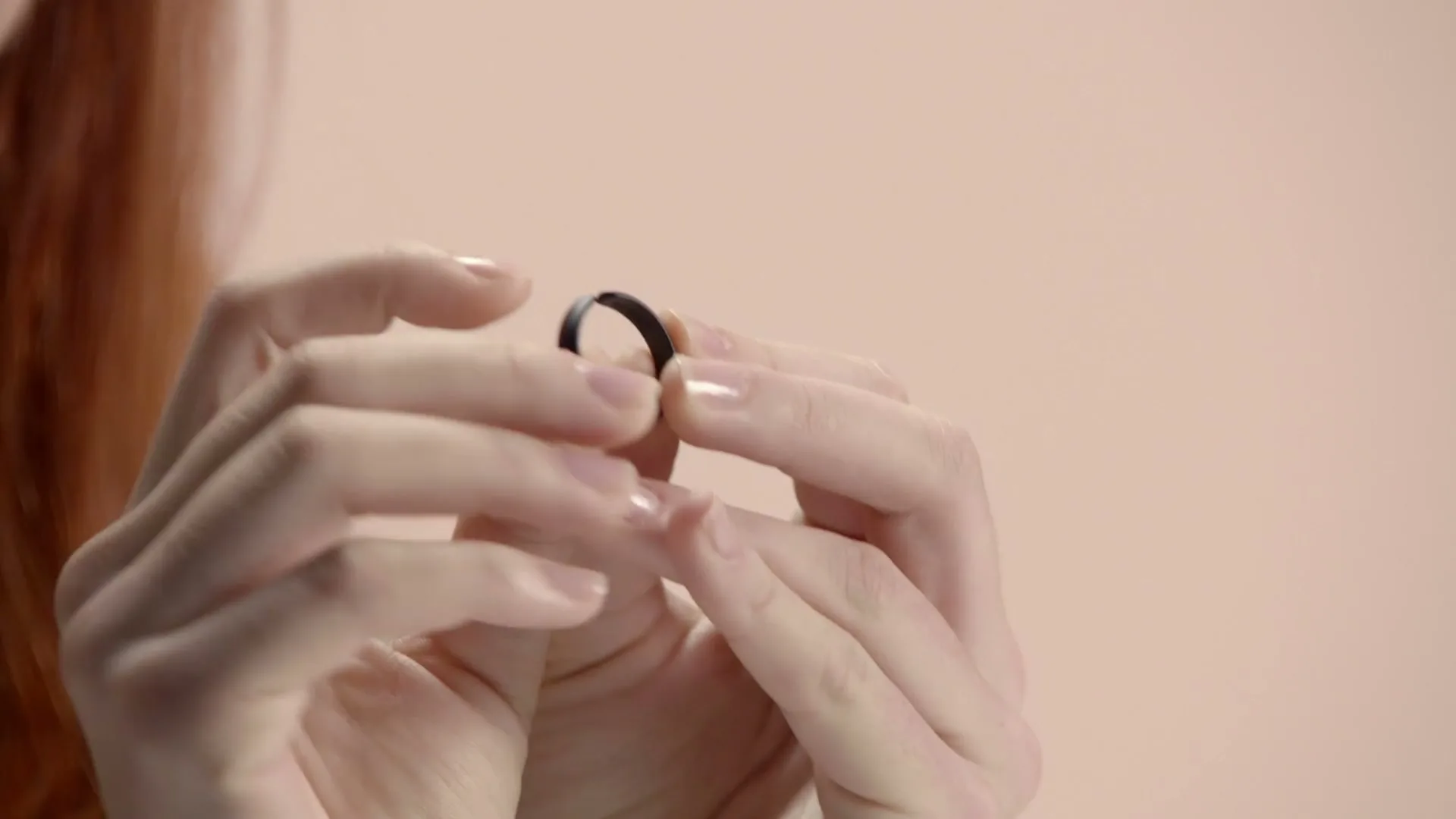 A woman with red hair holds a small black ring between her manicured fingers, examining it closely against a light pink background.